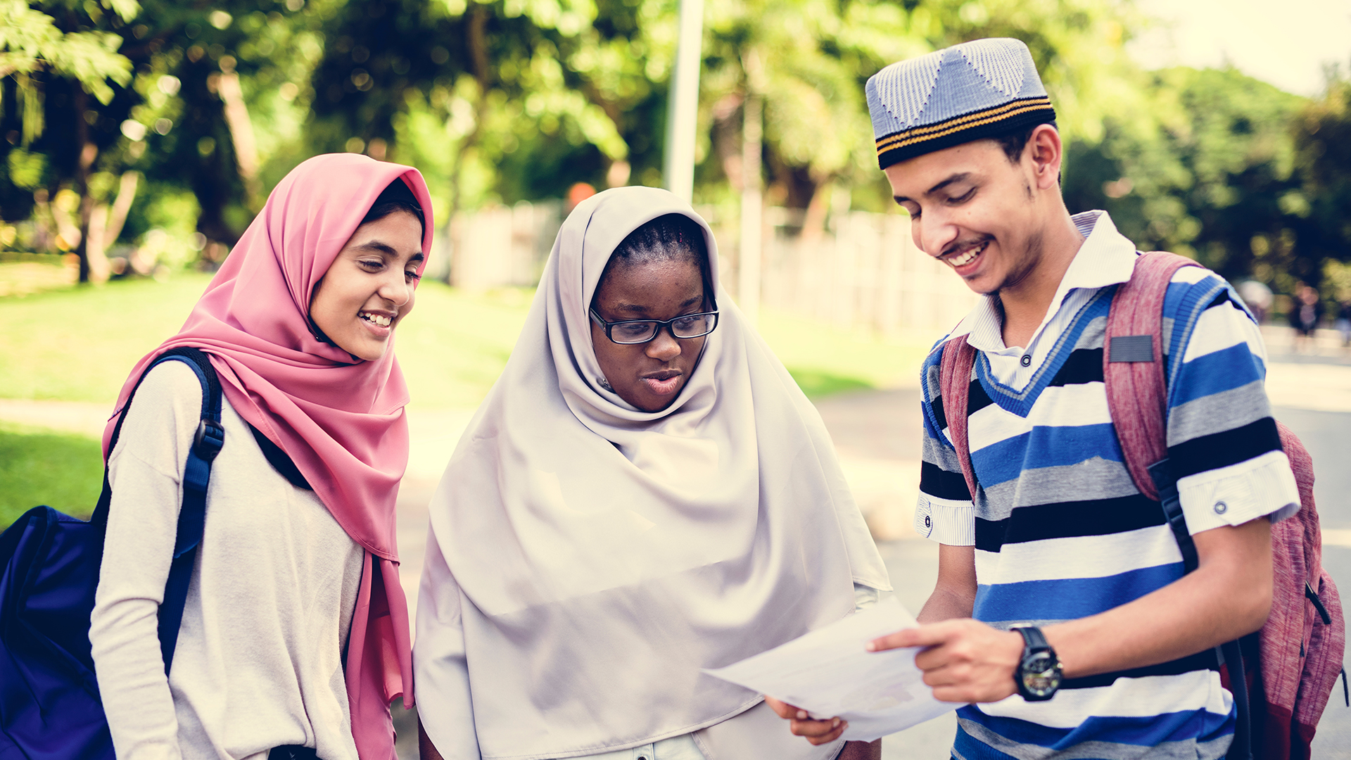 Photo of diverse group of two young women wearing headscarves and a young man wearing a hat looking at a paper.